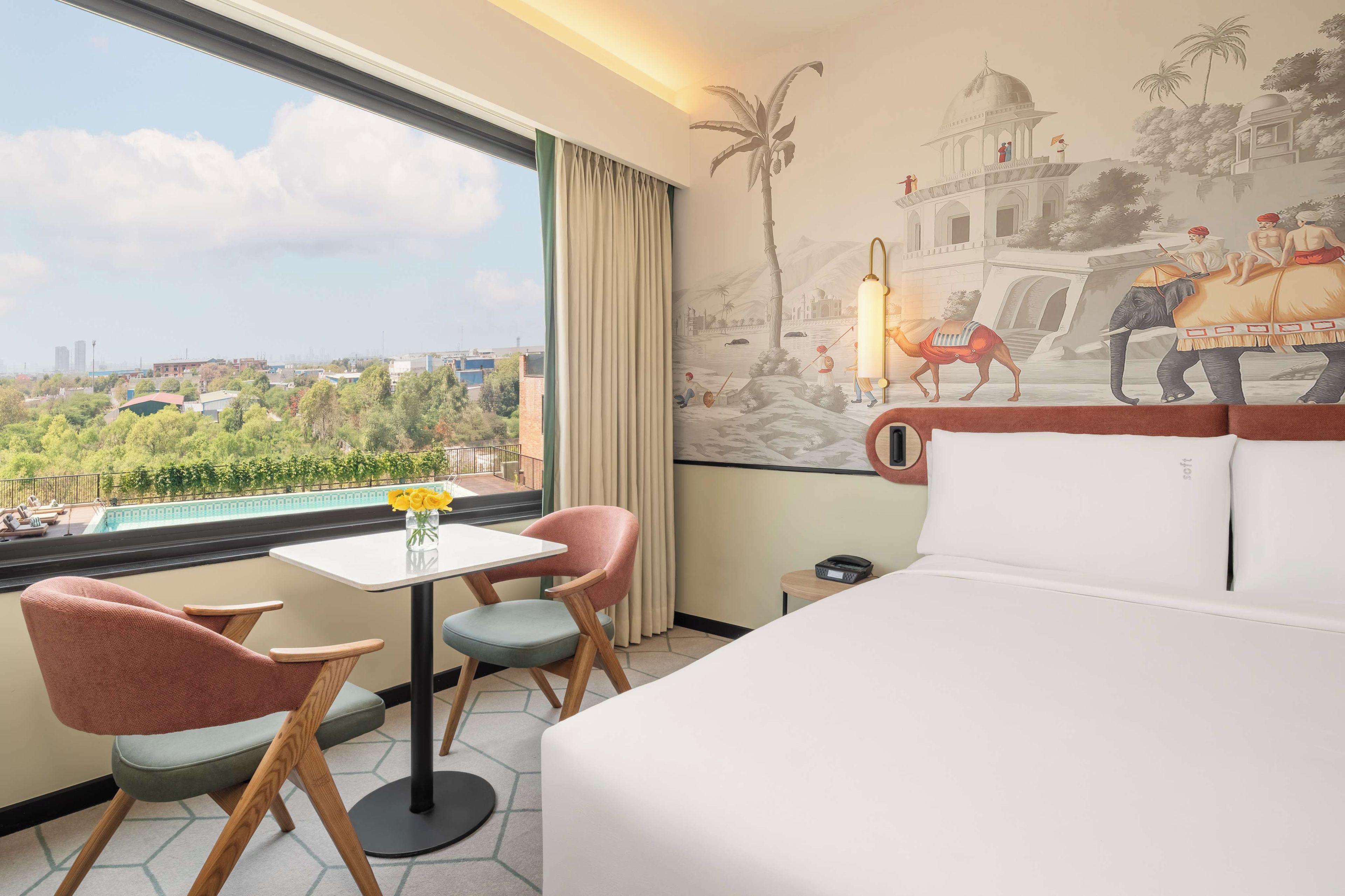 Une chambre d’hôtel avec un lit, deux chaises, une petite table sur laquelle est posé un vase de fleurs, une vue sur le paysage urbain et une fresque murale.