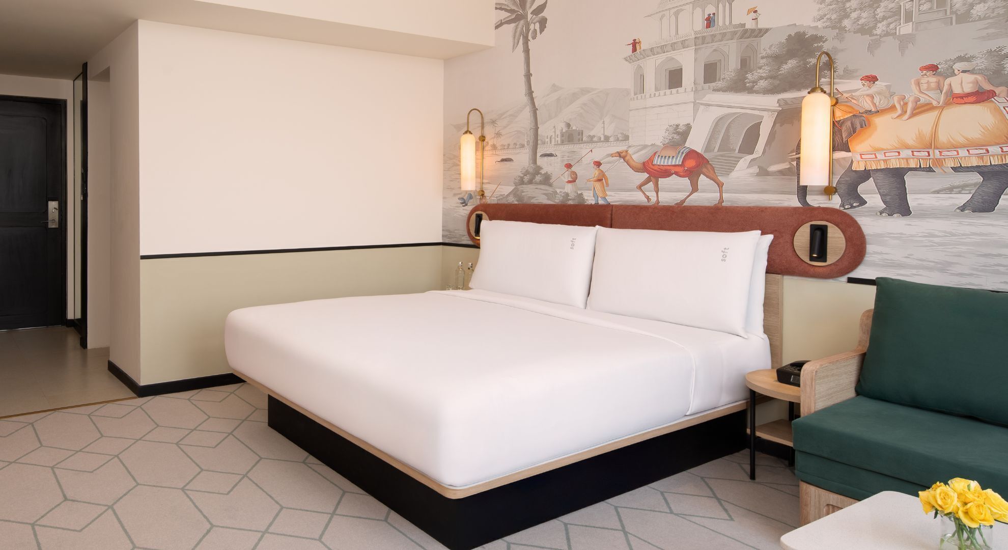 A hotel room with a large bed, a green armchair, a small table with a vase of flowers, and a mural depicting a historical scene.
