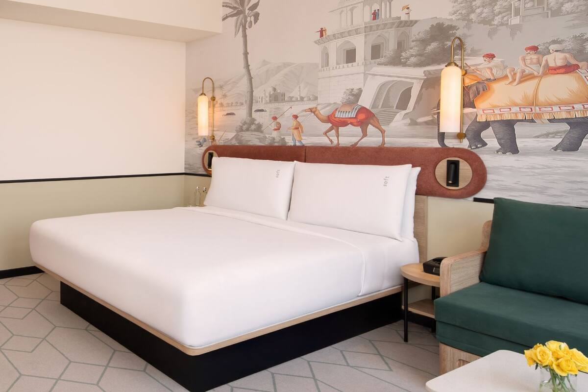 Una habitación de hotel con una cama grande, un sillón verde, una mesita con un florero y un mural que representa una escena histórica.