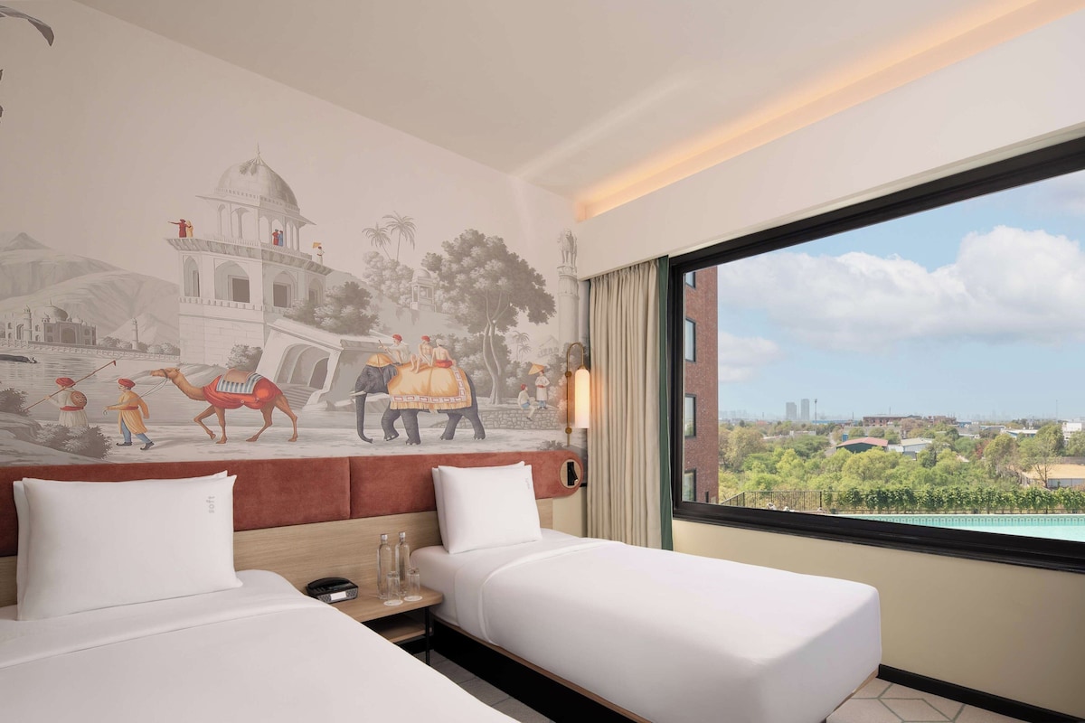 A hotel room with two beds, a mural of a historical scene, a view of the city, and a window with curtains.