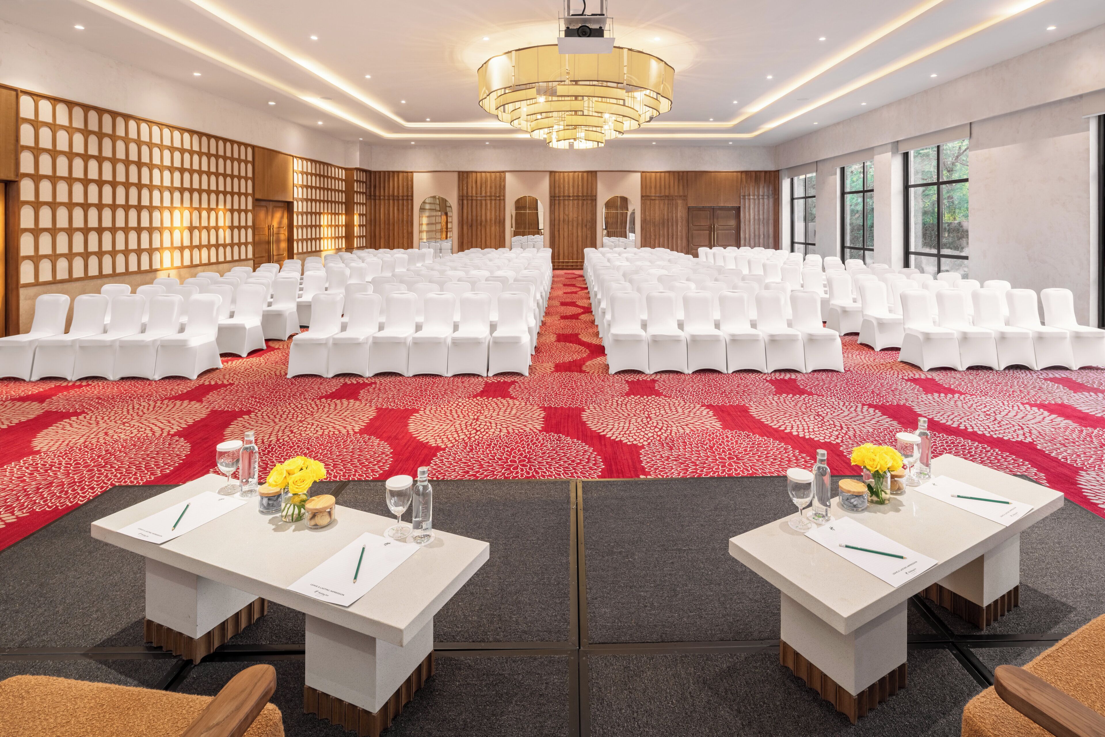 A conference room with rows of white chairs, a red patterned carpet, and two tables with name cards and pens.