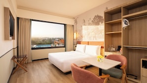 Habitación de hotel con cama, mesa de comedor, sillas, televisión y un gran ventanal con vistas.