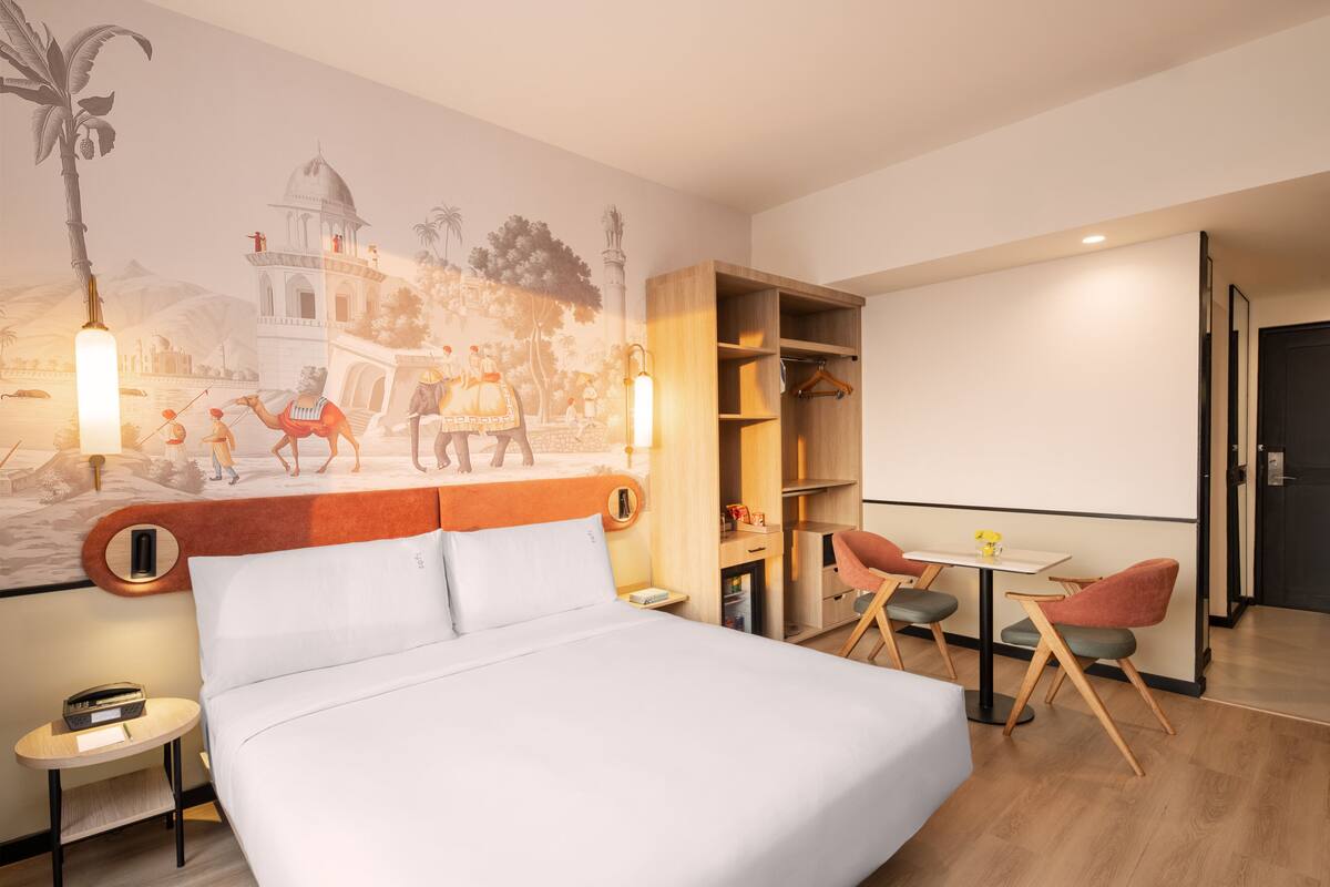 Ett hotellrum med en säng, ett litet bord, två stolar, en bokhylla och en väggmålning.