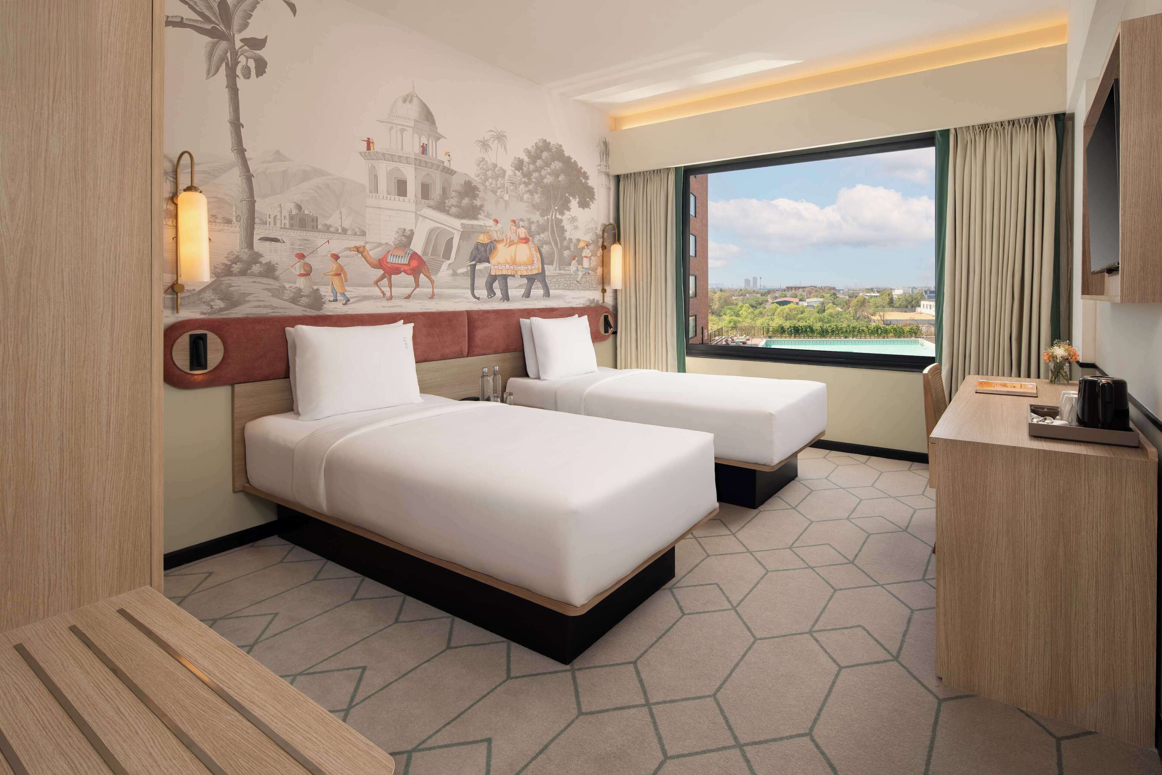 Habitación de hotel con dos camas, un ventanal con vistas, un mural en la pared y un escritorio con lámpara.