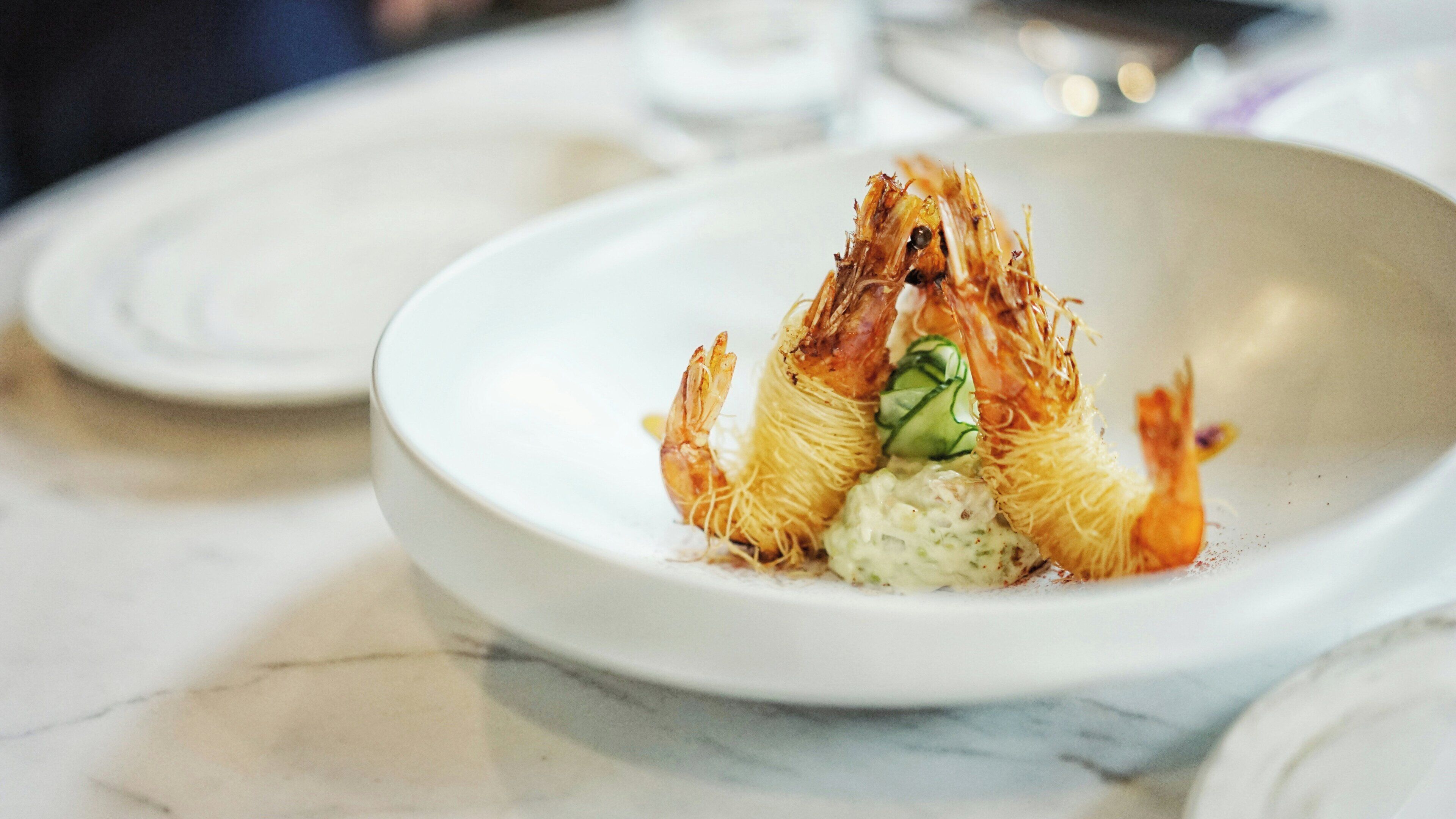 A white plate with a dish containing shrimp and a green vegetable, possibly a cucumber, on a marble surface.