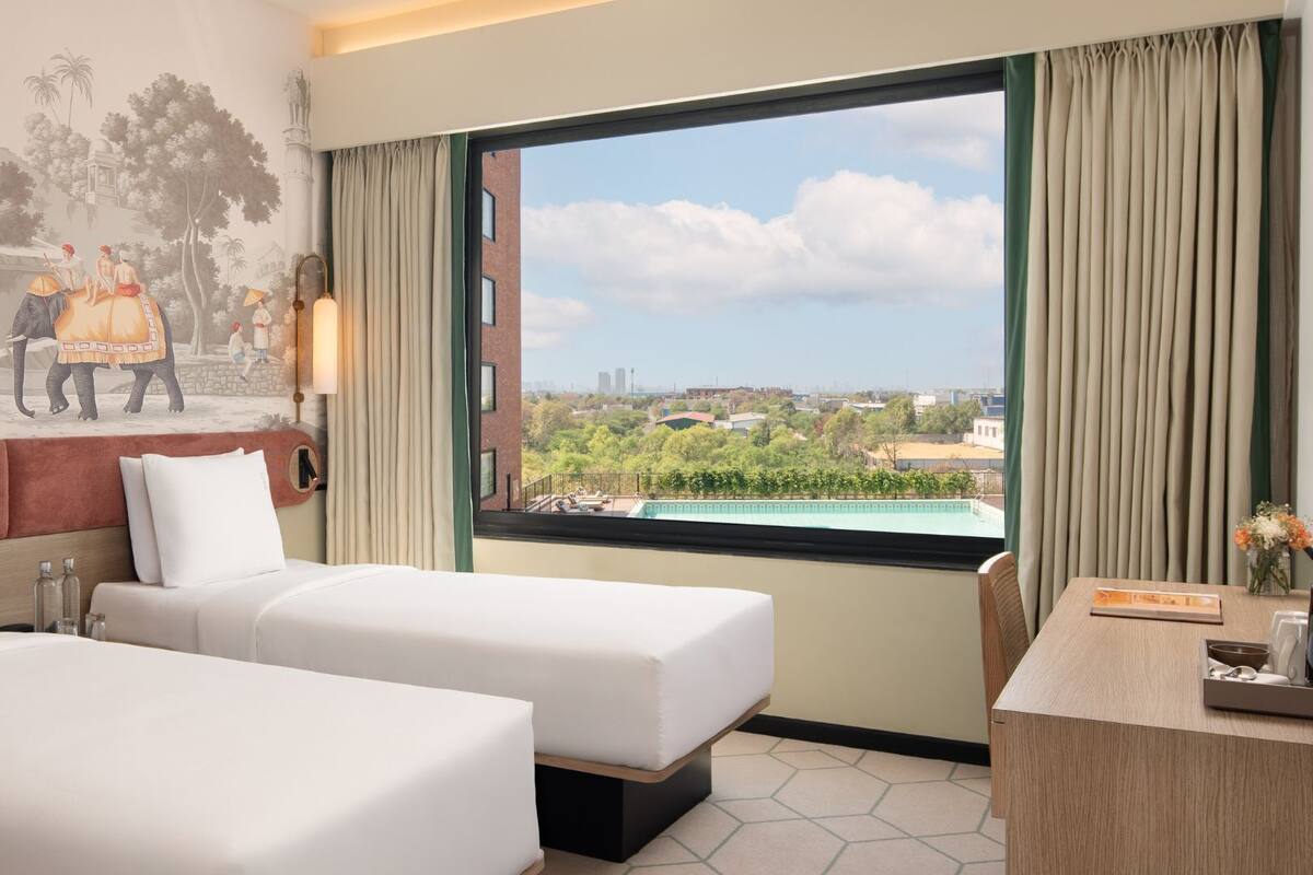 Habitación de hotel con dos camas, un mural que representa una escena histórica, un ventanal con vistas a la ciudad y un escritorio con un florero.