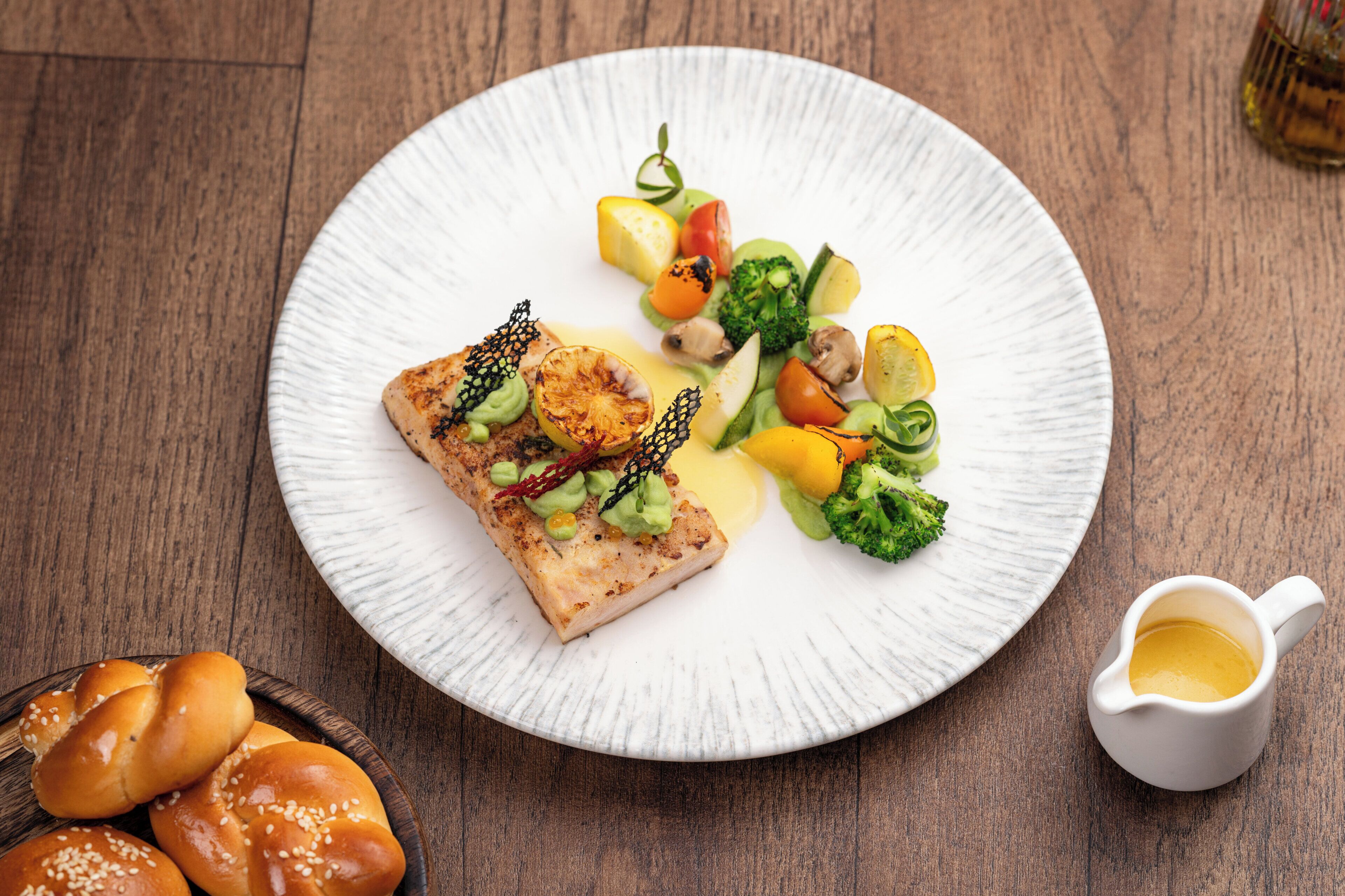 A plate of food with a piece of fish, vegetables, and a small jug of sauce.