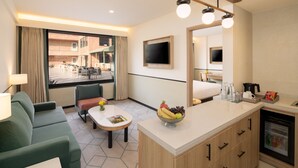 A hotel room with a sofa, a dining table, a kitchen area with a refrigerator, a TV mounted on the wall, and a large window offering a view of the outside. - Holiday Inn Gurgaon NH8 by IHG (Gurugram)