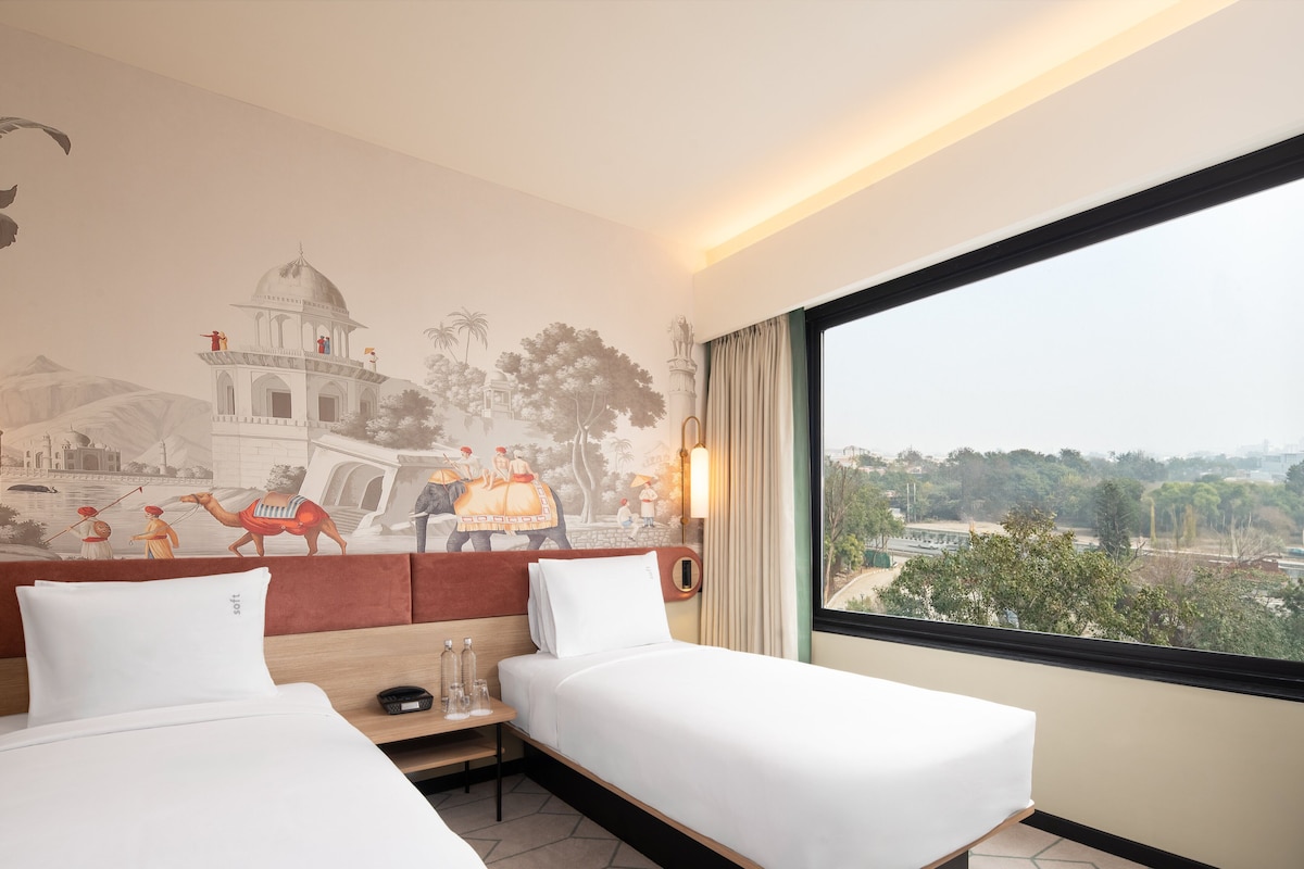 A hotel room with two beds, a mural on the wall, a window with a view, and a nightstand with a lamp.