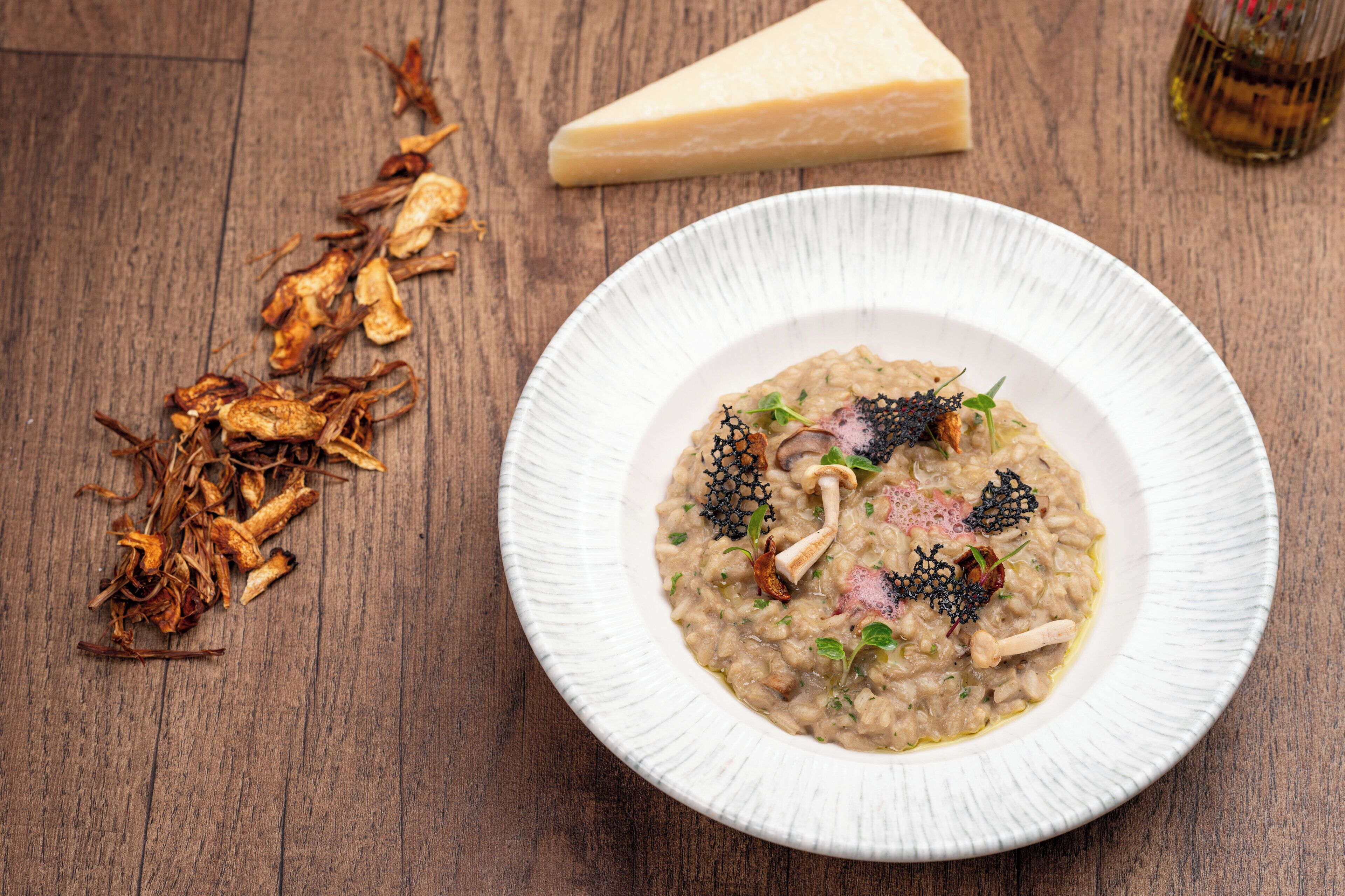 A plate of risotto with mushrooms and herbs, accompanied by dried mushrooms and a wedge of cheese on a wooden table.