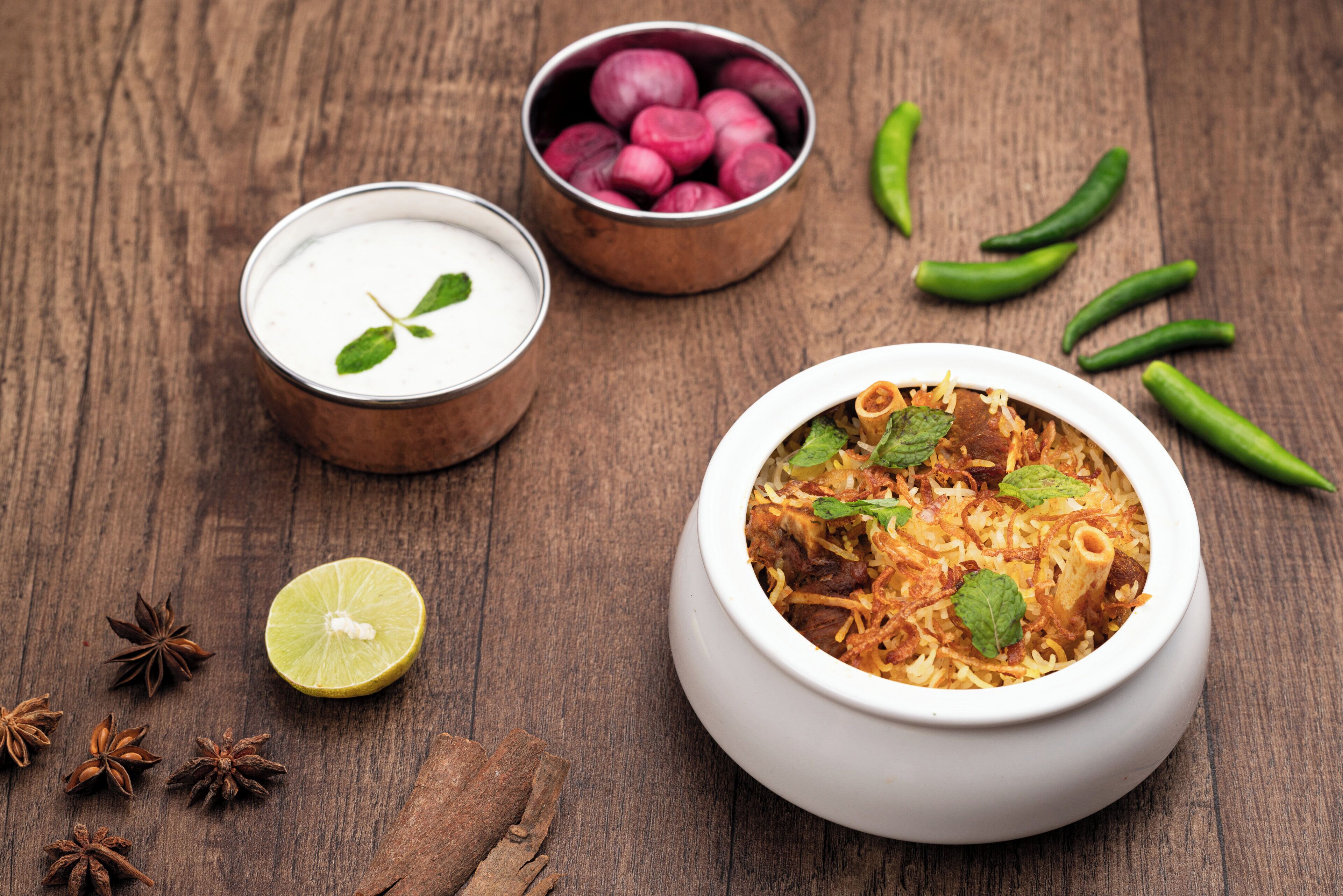 Un cuenco de biryani con hojas de menta, un cuenco de yogur y un cuenco de chiles rojos sobre una superficie de madera.
