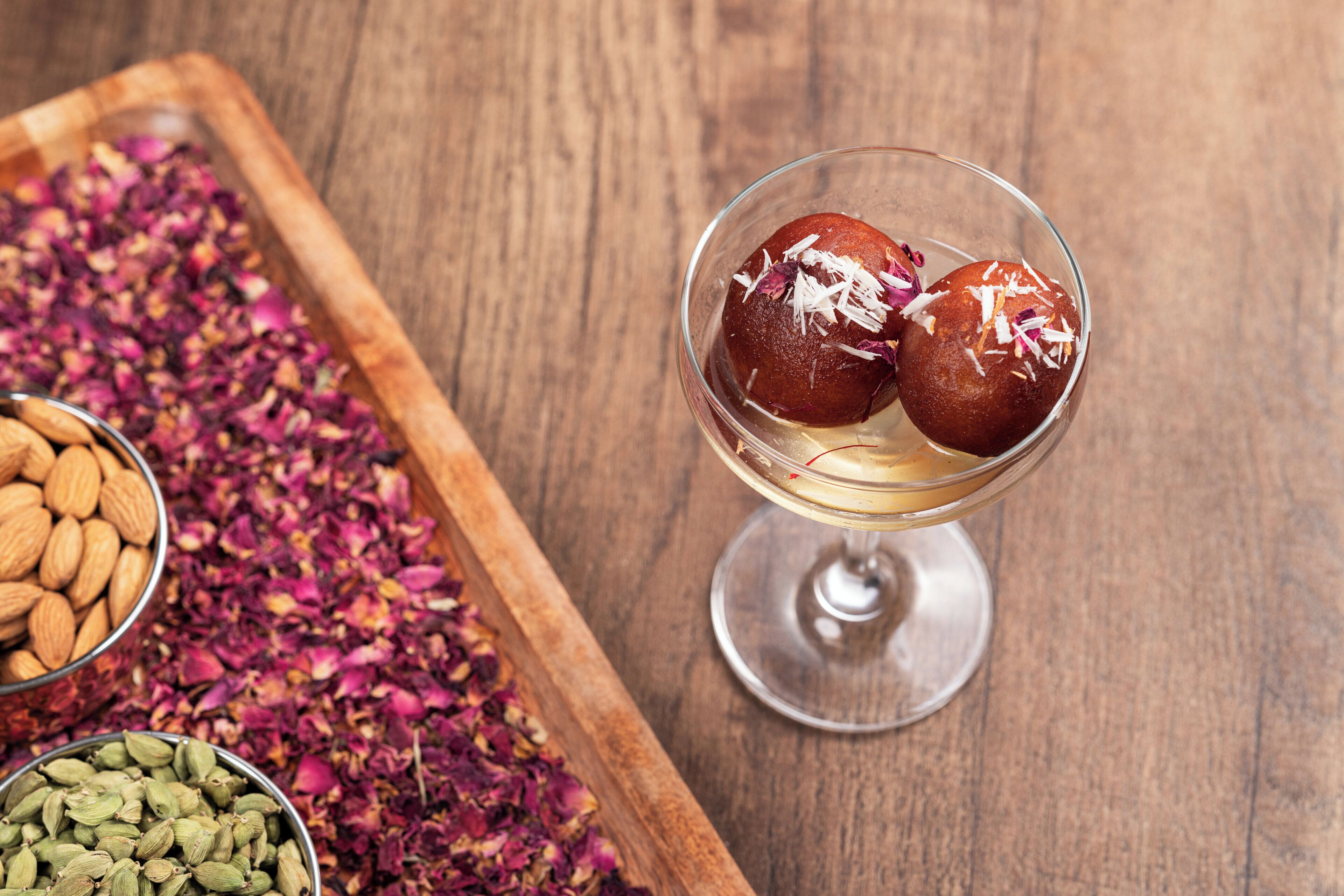 Ein Dessert mit zwei runden Süßigkeiten im Glas, serviert auf einer Holzschale mit Mandeln und getrockneten Blütenblättern.