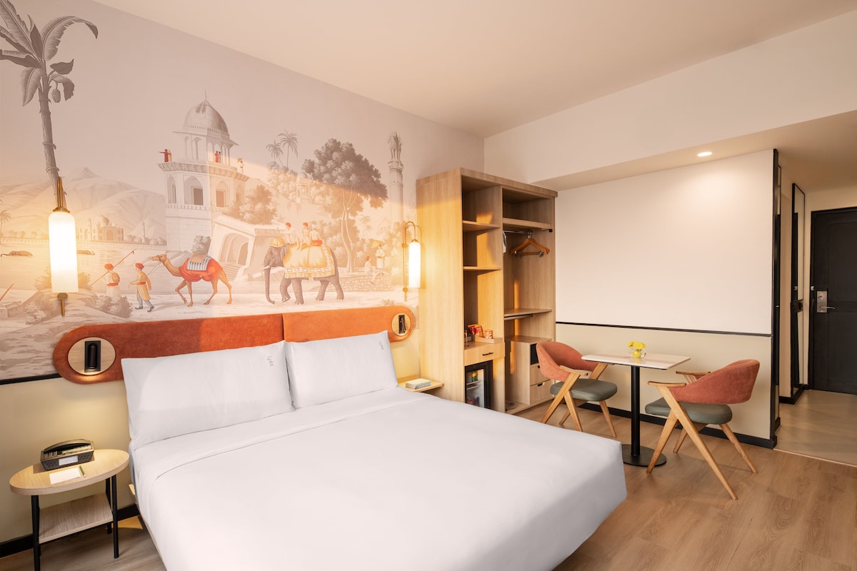 A hotel room with a bed, a small table, two chairs, a bookshelf, and a mural on the wall.