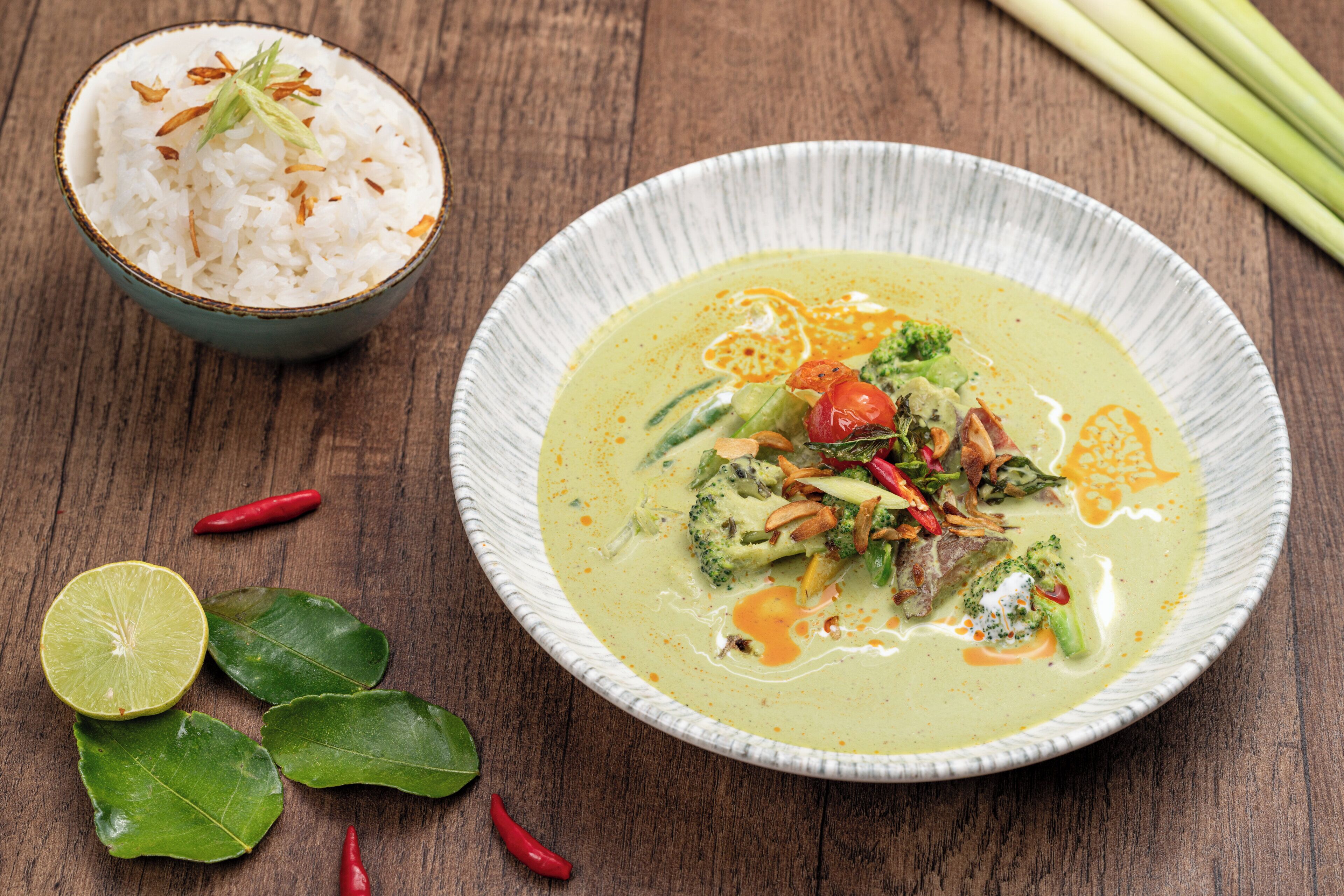 Un bol de curry vert aux légumes et à la viande, un bol de riz blanc, ainsi que quelques feuilles vertes et des piments rouges sur une table en bois.