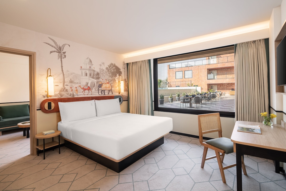 A hotel room with a bed, a desk, a chair, a small table, a lamp, a window with curtains, and a mural on the wall.