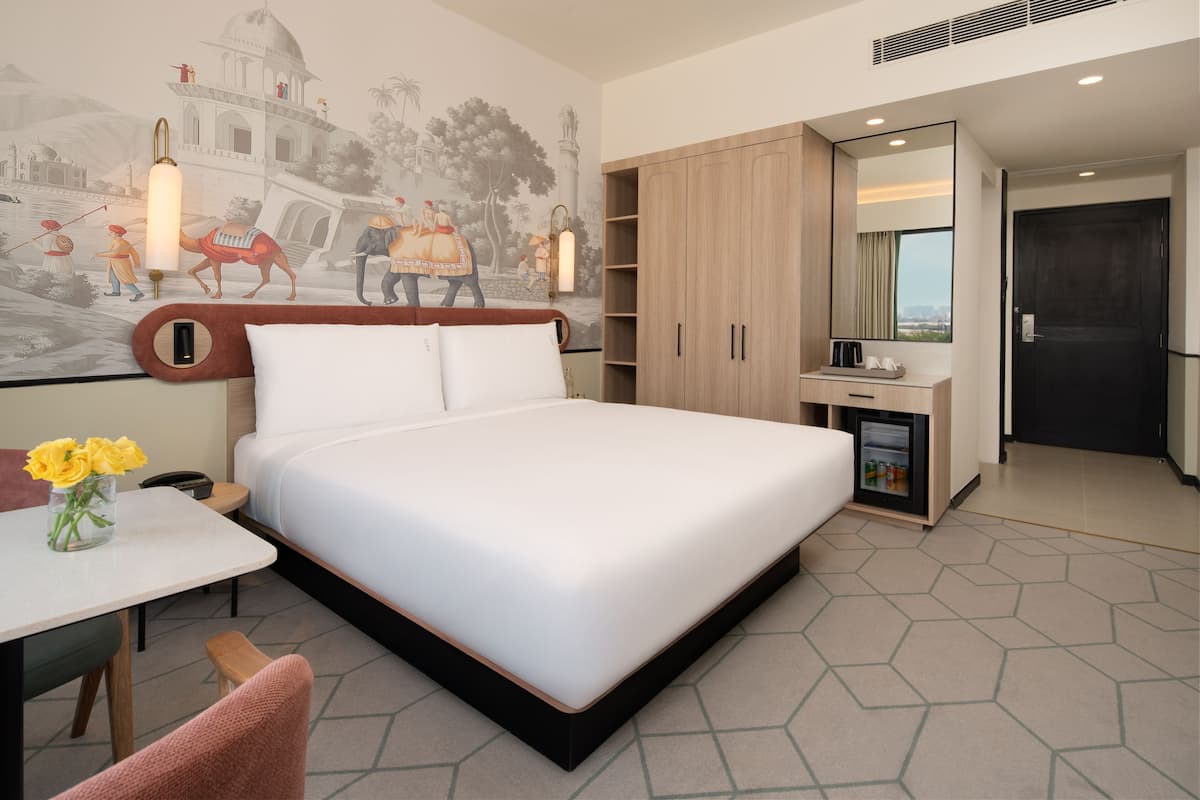 Habitación de hotel moderna con una cama grande, un escritorio pequeño con un florero y una minibar, además de un mural que representa una escena histórica.