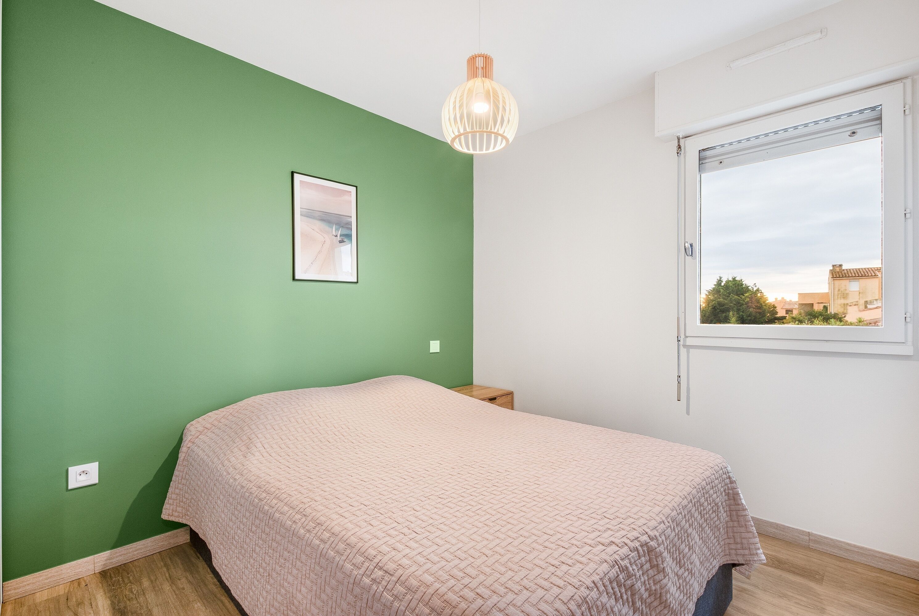 A bedroom with a green wall, a bed with a pink blanket, a wooden nightstand, a hanging light fixture, and a window with a view of buildings.