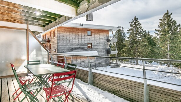 A wooden deck with a table and chairs, a metal railing, and a view of a building with a stone facade. - Joubarbes - Station à pied (Chamrousse)