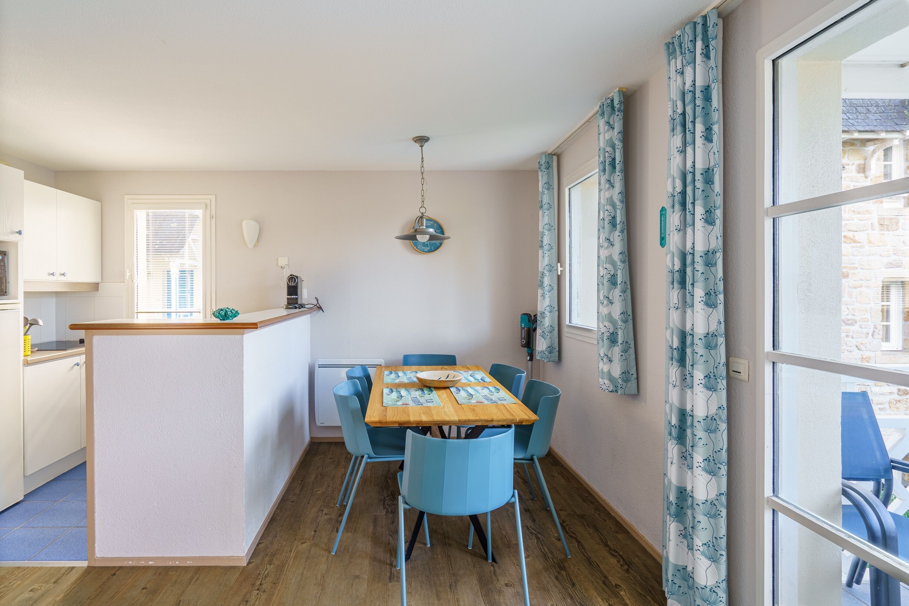 A compact kitchen with a dining area, featuring a wooden table, blue chairs, and a small island with a bowl.
