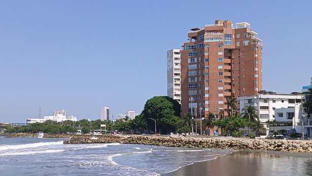 A coastal cityscape with a prominent multi-story building, a rocky shoreline, and palm trees.