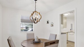 Un comedor con una mesa redonda, seis sillas, una araña, un ventanal con persianas y vistas a la cocina.