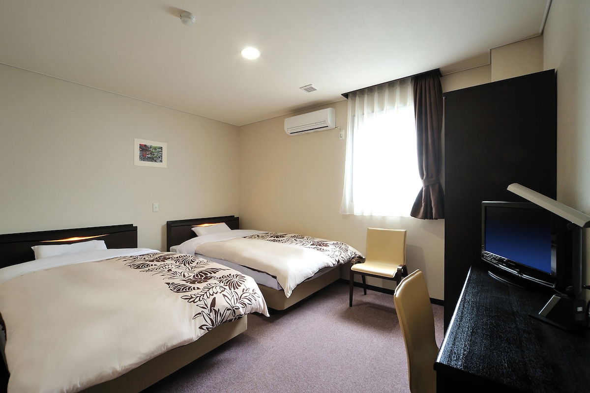 Une chambre d’hôtel avec deux lits, un bureau avec un ordinateur et une fenêtre avec des rideaux.