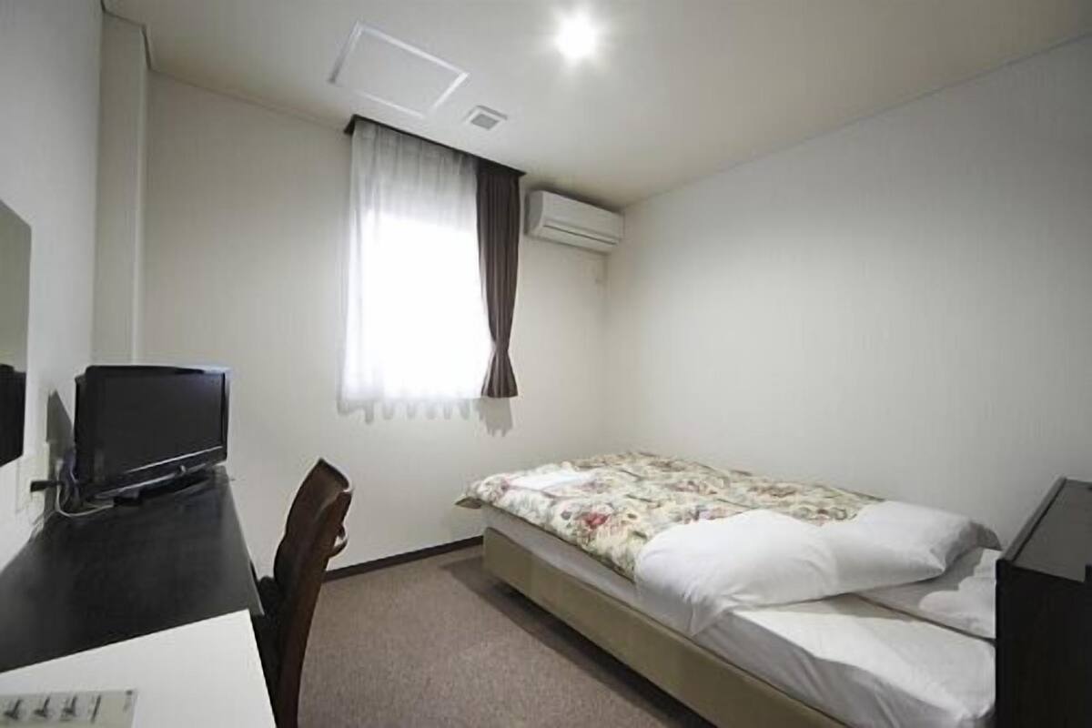 Une petite pièce, peu meublée, comprenant un lit, un bureau, une chaise et un téléviseur.
