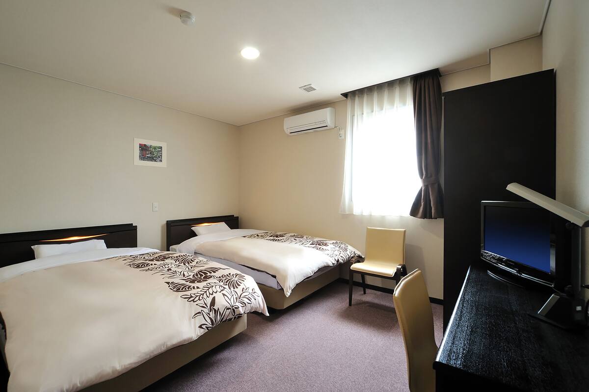 Une chambre d’hôtel avec deux lits, un bureau avec un ordinateur et une fenêtre avec des rideaux.