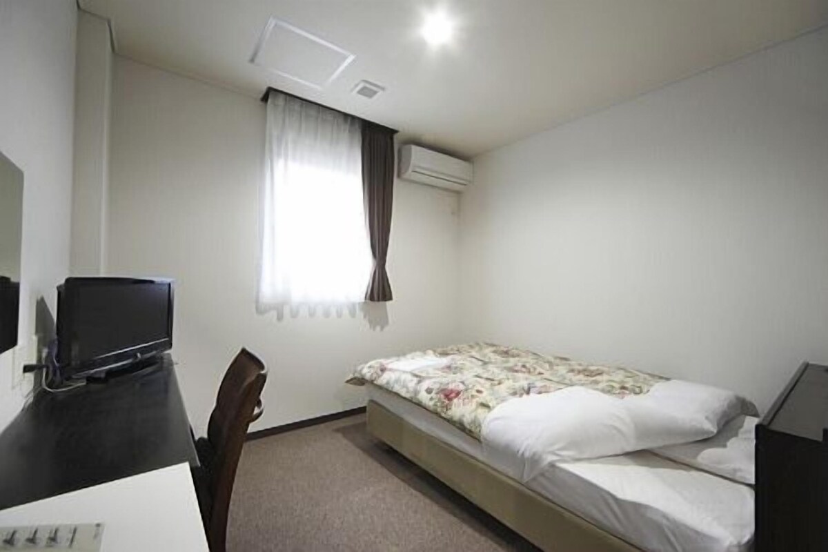Une petite pièce, peu meublée, comprenant un lit, un bureau, une chaise et un téléviseur.