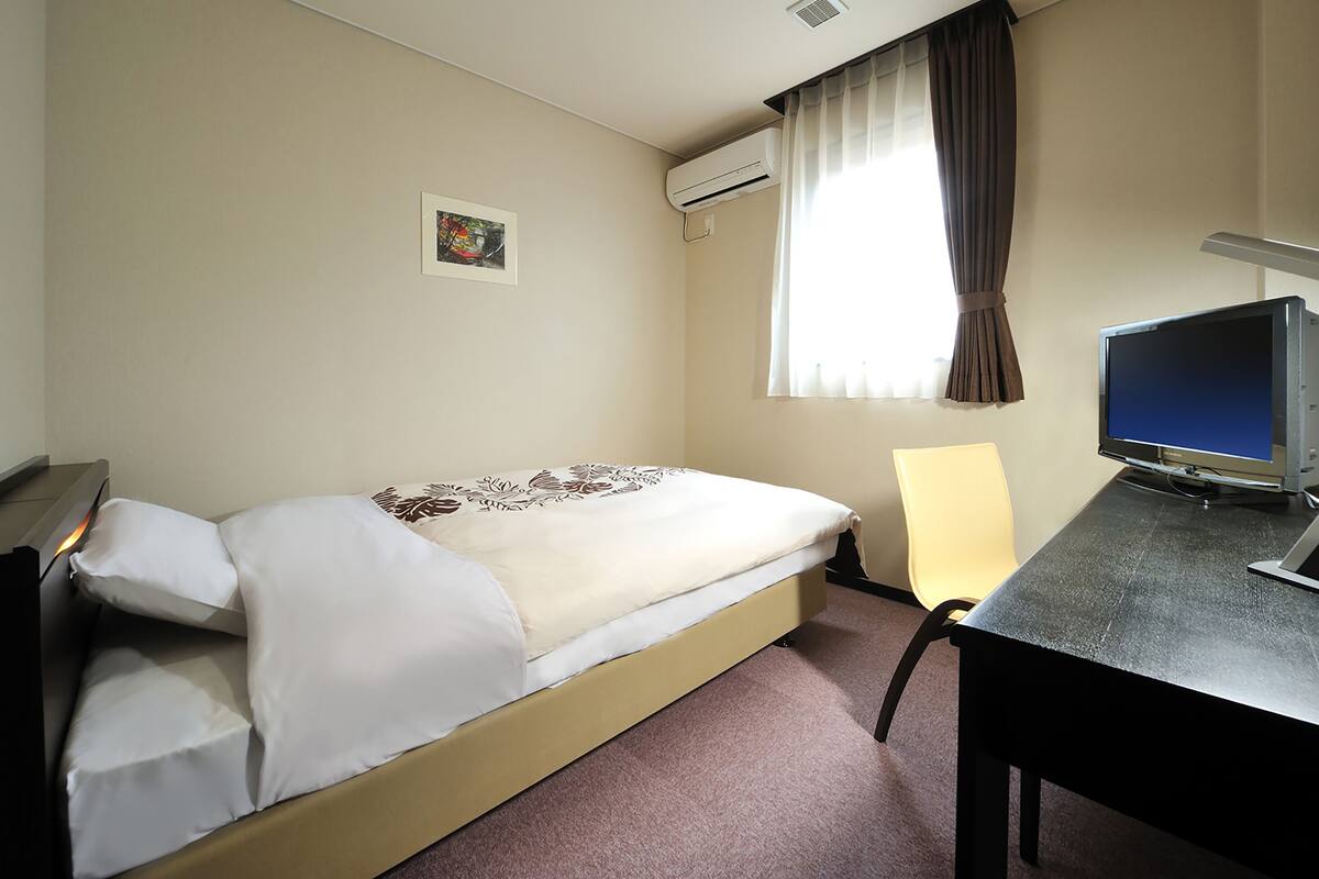 Une chambre d’hôtel avec un lit, un bureau avec un ordinateur, une chaise, un climatiseur, une fenêtre avec des rideaux et un petit tableau accroché au mur.
