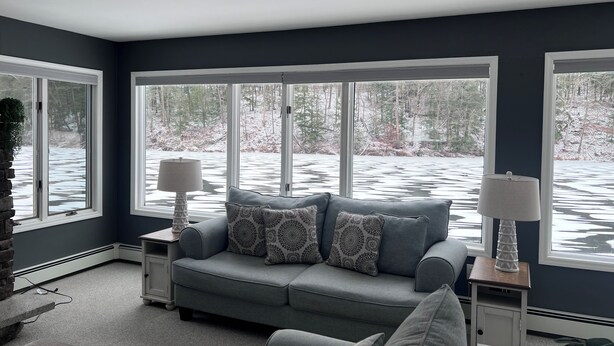 Un salón con un sofá gris, cojines estampados, una mesita de noche con una lámpara y ventanas amplias con vistas a un paisaje nevado.