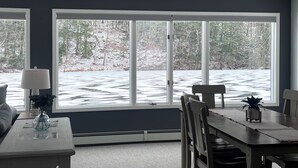 Un área de comedor con una mesa y sillas, ventanas amplias con vistas a un paisaje nevado y una lámpara sobre una mesita auxiliar.