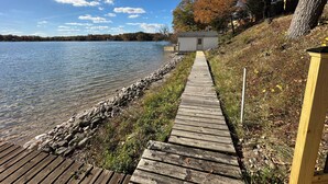 A wooden dock leading to a small white building by a lake. - Pine Lake Cottage with Views (Plainwell)