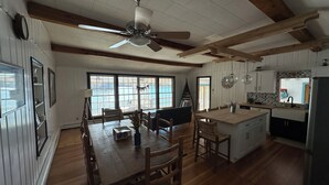 A spacious kitchen with wooden floors, a dining area with a large table and chairs, a ceiling fan, and a view through the windows. - Pine Lake Cottage with Views (Plainwell)