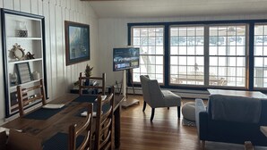 A cozy living room with a wooden dining table, chairs, a TV, and a large window offering a view of a snowy landscape. - Pine Lake Cottage with Views (Plainwell)