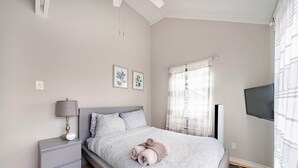 A bedroom with a bed, bedside table, lamp, wall art, window with curtains, and a TV.