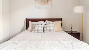 Ein ordentlich bezogenes Bett mit weißer Daunendecke und zwei karierten Kissen, ein Nachttisch mit Lampe sowie ein an der Wand hängendes abstraktes Gemälde.