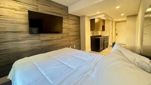 Une chambre d’hôtel moderne dotée d’un grand lit, d’un téléviseur à écran plat fixé à un mur en bois, d’une petite kitchenette et d’un dressing.