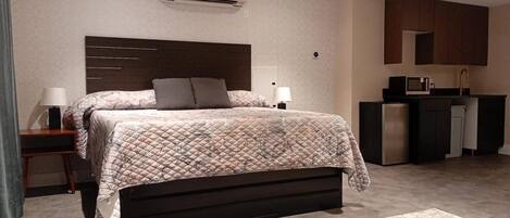 A modern bedroom with a large bed, bedside lamps, a built-in desk, a kitchenette, and an air conditioning unit.