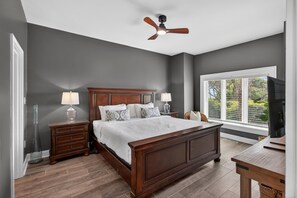 A bedroom with a wooden bed, nightstands, a ceiling fan, a window with blinds, and a TV. - Exclusive Seascape in West Beach Village! 3 King Beds! Oceanfront! (Kiawah Island)