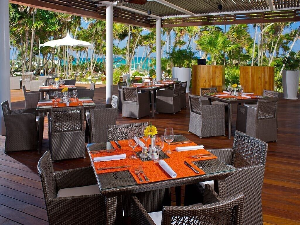 Un espace extérieur aménagé pour les repas, avec des meubles en osier et des sets de table orange, entouré de palmiers et offrant une vue sur l’océan.