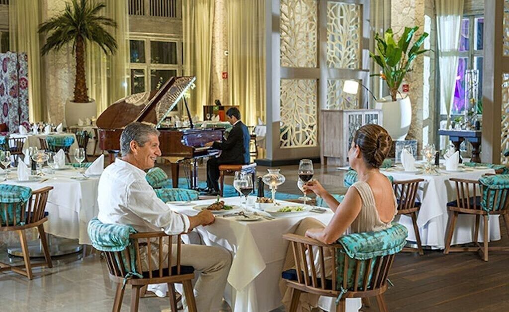 Une salle à manger élégante, avec des tables dressées pour deux personnes, un grand piano et un pianiste.