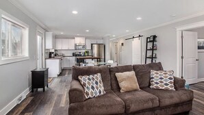 A modern living room with a brown sectional sofa, a kitchen in the background, and a hallway leading to a door. - Sandcastle Muskegon Beach Cottage (Muskegon)