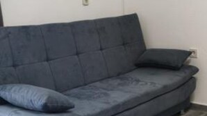 Un sofá gris con un cojín, apoyado contra una pared lisa donde se ve un enchufe.