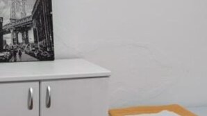 Un mueble blanco con tiradores plateados, una fotografía en blanco y negro de una escena callejera y una pequeña mesa con un paño blanco doblado y un libro.