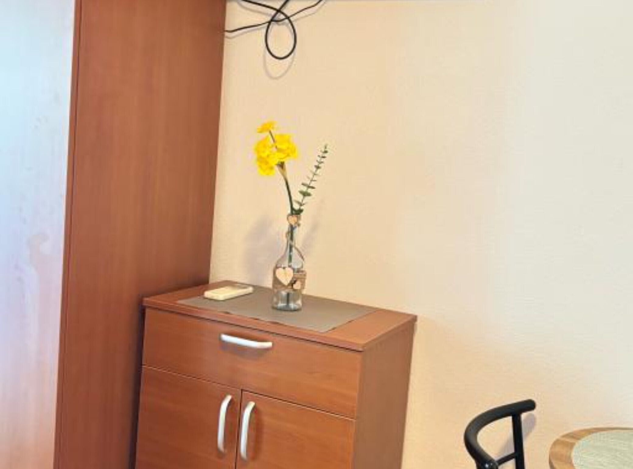 Un mueble de madera con un jarrón que contiene una flor amarilla, un teléfono inteligente encima y una silla al fondo.