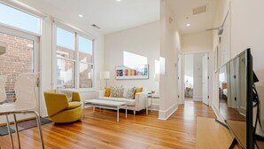 A spacious living room with a sofa, yellow armchair, and a coffee table. There is a TV on a wooden stand and a large window with a view of the outside. - The Daily Condo 201 at ATB (Asheville)