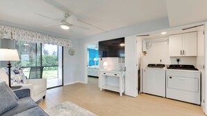 Una sala moderna con un sofá gris, una mesa de centro y una lámpara. Cuenta con una zona de cocina con estufa, microondas y armarios. La habitación tiene ventanas amplias y un ventilador de techo.