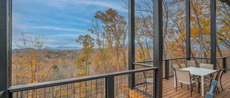 A wooden deck with a table and chairs, overlooking a forest with autumn foliage.