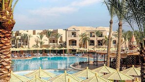 A pool area with lounge chairs and umbrellas, surrounded by palm trees and a building with a Mediterranean architectural style. - Magic World Sharm - Club By Jaz (Sharm El Sheikh)